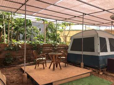 Un aménagement extérieur confortable comprend une tente, des meubles en bambou sur une plateforme en bois, des plantes vertes luxuriantes et un toit ombragé, créant ainsi un espace paisible inspiré de la nature.