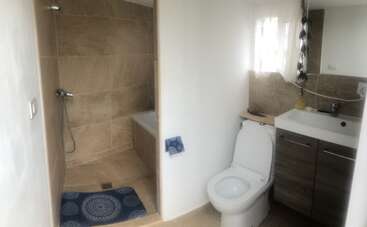 Cette salle de bain comprend une douche à l'italienne avec du carrelage marron, un tapis bleu, des toilettes, un lavabo moderne avec rangement et une petite fenêtre avec des rideaux transparents.