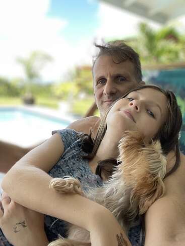 Um homem e uma menina estão tirando uma selfie aconchegante à beira da piscina, com um cachorrinho aninhado nos braços da menina, ao ar livre.