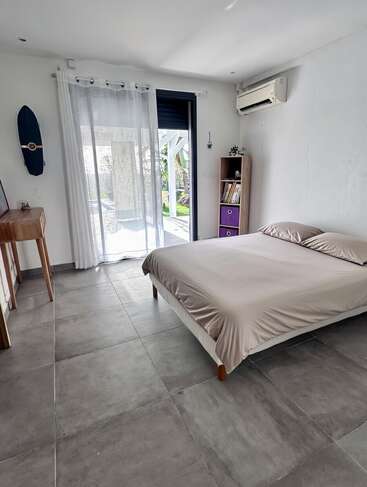 Este é um quarto bem iluminado e minimalista, com piso de azulejos cinza, cama de casal, escrivaninha de madeira, estante, ar-condicionado, cortinas transparentes e porta de correr que dá para o exterior.
