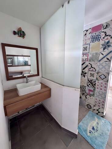 Uma casa de banho moderna conta com um lavatório de madeira com pia tipo bacia, um espelho grande, uma obra de arte decorativa com peixes, uma divisória de vidro fosco, azulejos estampados no chuveiro e um tapete azul com tema de peixes.