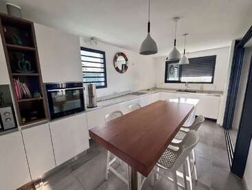 Esta cozinha moderna tem armários brancos minimalistas, uma ilha de madeira com bancos de bar brancos, duas janelas grandes, luminárias suspensas, prateleiras abertas, forno embutido e elementos de decoração elegantes.