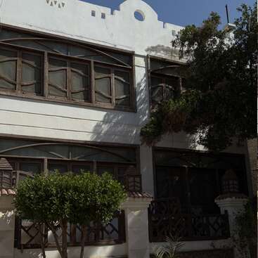 The image depicts a white building with brown-framed windows and a balcony, featuring trees and a blue sky in the background on a sunny day.