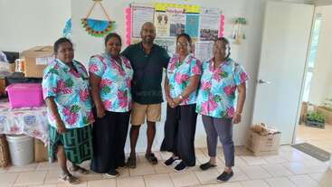 Fünf Personen posieren in einem hellen Raum, vier Frauen in passenden geblümten Hemden und ein Mann in Freizeitkleidung, die vor einem schwarzen Brett stehen.