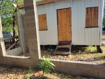 Eine kleine Wellblechhütte mit Holztür und Fensterverkleidung steht hinter einer unfertigen Blockwand, umgeben von Bäumen und trockenem Gras im Sonnenlicht.