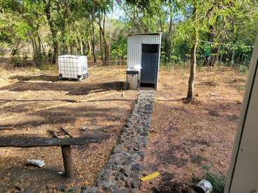 Ein kleiner Außenbereich mit einem einfachen Metallschuppen, einem Wassertank, einem Steinweg, verstreutem Geröll, Bäumen und trockenem Boden lässt auf eine einfache ländliche oder gärtnerische Umgebung schließen.