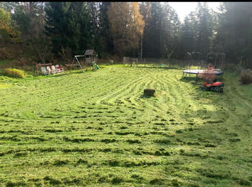 Ein großer, frisch gemähter Garten mit spielerischen Rasenmäherspuren. Auf der sonnigen, von Bäumen umgebenen Lichtung stehen Schaukeln, eine Rutsche, Trampoline und Stühle.