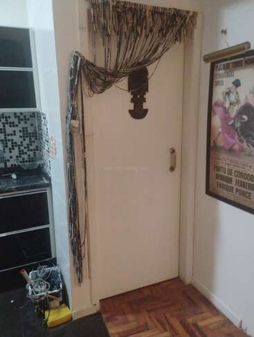 A white door decorated with hanging beads, wall art, and a wooden handle. To the left, a kitchen area with black tile and cleaning supplies is visible.