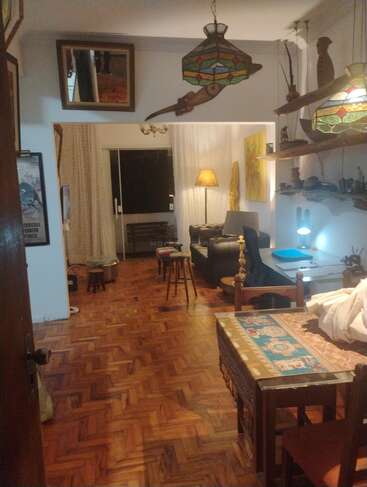 A cozy living room with wooden parquet floor, vintage decor, hanging stained glass lamps, a dining table with tapestry, desk, floor lamp, stools, and eclectic art.