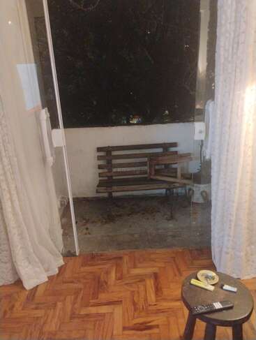 A cozy room with parquet flooring opens to a small balcony featuring a wooden bench. Sheer curtains frame glass doors. A black stool holds various items.