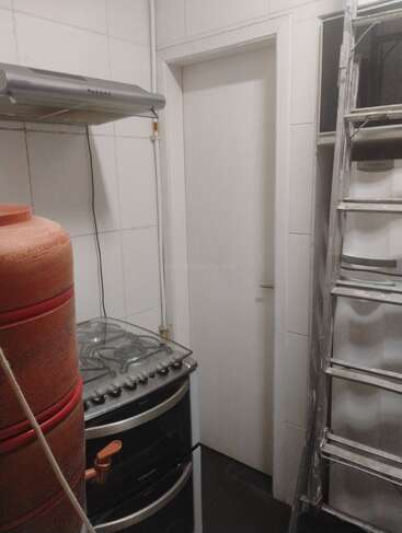 This image shows a small kitchen corner with a stove, red water dispenser, a metallic ladder, white tiled walls, and a closed white door.