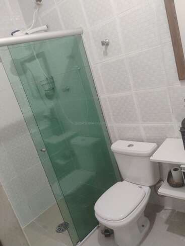 This is a small bathroom with white tiled walls, a toilet, a glass shower enclosure, a shelf, toiletries, and a partial view of a mirror.