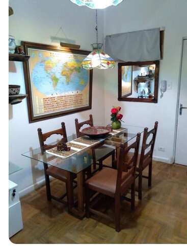 This cozy dining area features a glass table with four wooden chairs, a world map, decorative pendant lamp, mirror, shelves, and a vase of flowers.