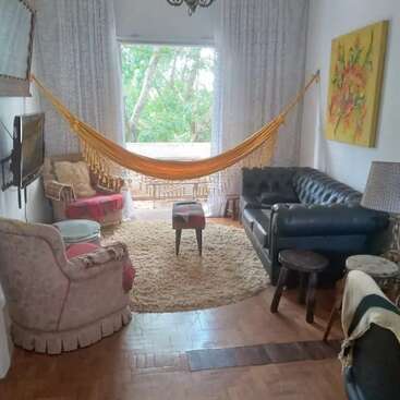 Cozy living room with a yellow hammock, round rug, comfortable chairs, black leather sofa, TV, side tables, window with tree view, colorful wall art.