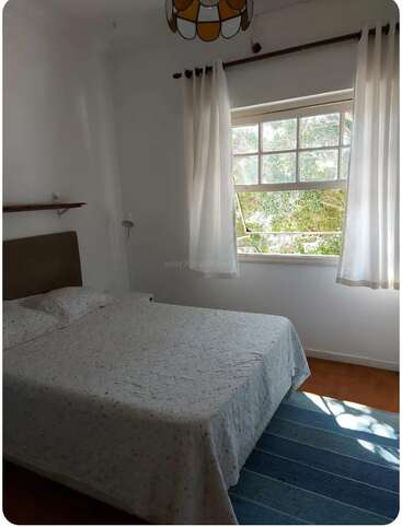 A bright, cozy bedroom with a double bed, white bedding, and curtains. Sunlight streams through a large window, illuminating a blue rug on wooden floors.
