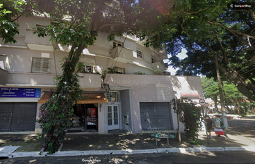 This image shows a street with a building that houses small shops, trees casting shade, and a sidewalk with two people walking and an abandoned shopping cart.