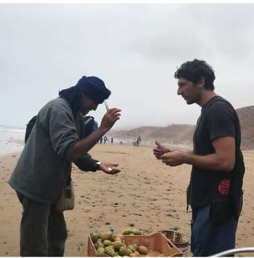 Deux hommes se tiennent sur une plage brumeuse, l'un vendant des figues de Barbarie à partir d'une caisse, tandis que l'autre inspecte ou achète le fruit pendant leur interaction.
