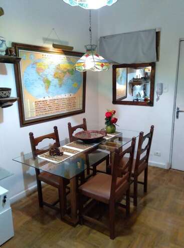 Ce coin repas confortable comprend une table en verre avec quatre chaises en bois, une carte du monde, une lampe suspendue décorative, un miroir, des étagères et un vase de fleurs.