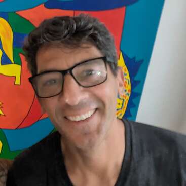 Un hombre con cabello oscuro y gafas sonríe a la cámara. Lleva una camisa oscura. Detrás de él hay un cuadro abstracto de colores vivos.