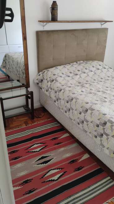 Une chambre confortable avec une tête de lit beige, un couvre-lit à motifs, un tapis à motifs rouges, une étagère en bois avec une cruche et un grand miroir reflétant une partie de la pièce.