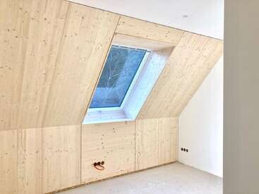 Une pièce en cours de construction présente des panneaux muraux en bois clair, une grande fenêtre inclinée, un sol non fini et des prises électriques apparentes, créant ainsi une atmosphère lumineuse, moderne et minimaliste.