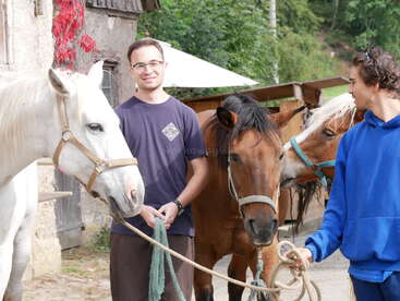 Dois jovens estão do lado de fora com três cavalos. Um homem segura uma corda presa a um cavalo branco, enquanto o outro usa um capuz azul, sorrindo.