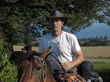 Ein Typ mit Cowboyhut und weißem T-Shirt reitet draußen auf einem braunen Pferd, im sonnigen Hintergrund sieht man Bäume und eine schöne Landschaft.