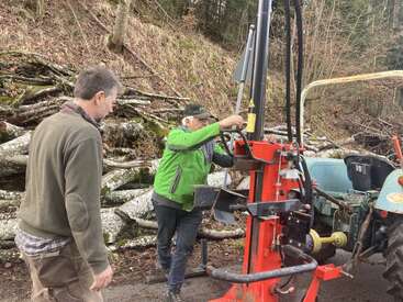 Two men are operating a log splitter machine beside a tractor. Behind them, there is a pile of cut logs in a forested, outdoor area.