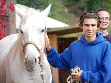 A young man in a blue hoodie gently pets a white horse while holding a rope. Another smiling man stands in the background, outdoors near a wooden building.