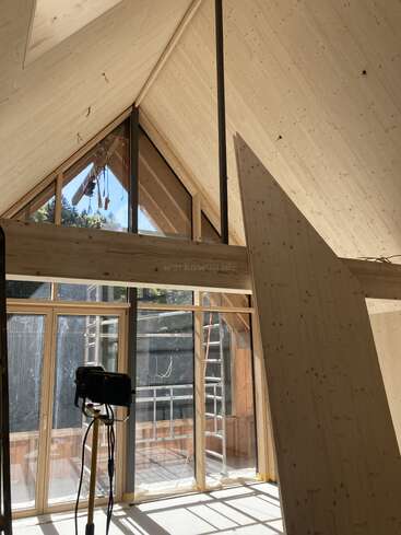 Esta imagem mostra um interior de madeira parcialmente construído, com grandes janelas triangulares, um teto alto e inclinado, a luz do sol entrando, uma câmera em um tripé e andaimes visíveis do lado de fora.