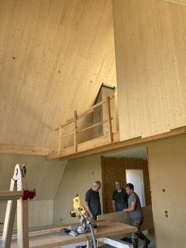 Três homens trabalham juntos dentro de um ambiente moderno com acabamento em madeira, ainda em construção. Ferramentas, uma escada e superfícies inacabadas indicam que a obra está em andamento. Acima, dá para ver um loft com corrimão.