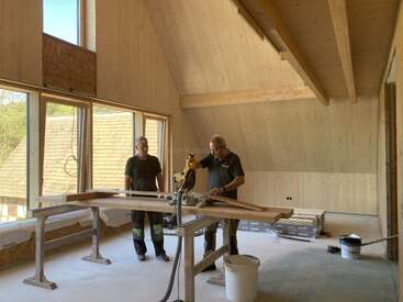 Dois homens estão trabalhando com madeira dentro de uma sala moderna, ainda em obras. Janelas amplas deixam a luz do sol entrar. Ferramentas, máquinas e materiais de construção estão espalhados por todo o espaço.