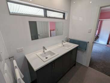 Salle de bains moderne avec double lavabo, grand miroir et armoires épurées. Comptoirs blancs, porte-serviettes et serviette bleue. Petite fenêtre au-dessus du miroir. La chambre adjacente aux murs roses est visible par la porte ouverte.