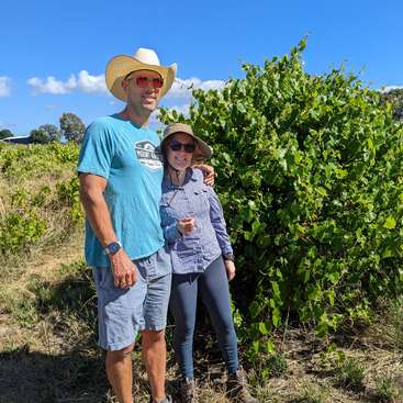 The image depicts a man and woman standing together in front of a lush green bush, with the man wearing a cowboy hat and the woman sporting a wide-brimmed sun hat.