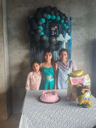 Drei Personen, wahrscheinlich eine Mutter und zwei Kinder, stehen hinter einem Tisch mit einem rosa Kuchen. Zur Dekoration gehören Luftballons, Geschenke und eine festliche Kulisse, die auf eine Feier schließen lässt.