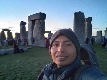 Ein Mann in warmer Kleidung lächelt für ein Selfie bei Stonehenge während des Sonnenuntergangs. Im Hintergrund sind die alte Steinstruktur und andere Besucher zu sehen.