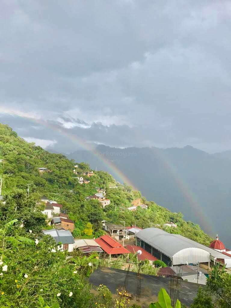 Ein üppig grünes Bergdorf unter einem dramatisch bewölkten Himmel mit bunten Dächern, lebendiger Vegetation und einem wunderschönen doppelten Regenbogen, der sich über der friedlichen Berglandschaft spannt.