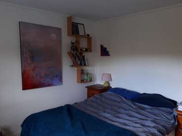 Um quarto aconchegante com uma cama de casal, roupa de cama azul e cinza, mesinhas de cabeceira de madeira, prateleiras de parede modernas, uma luminária suave, obras de arte abstratas e um ambiente tranquilo e minimalista.