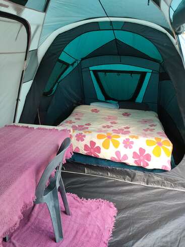 Inside a spacious tent, there’s a bed with bright floral sheets, a table covered with a pink cloth, a plastic chair, and a matching rug. Cozy setup.