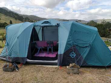 A large blue camping tent is set up outdoors, with its entrance open. Inside, there is a table, chairs, and a pink mat. Beautiful hills surround.