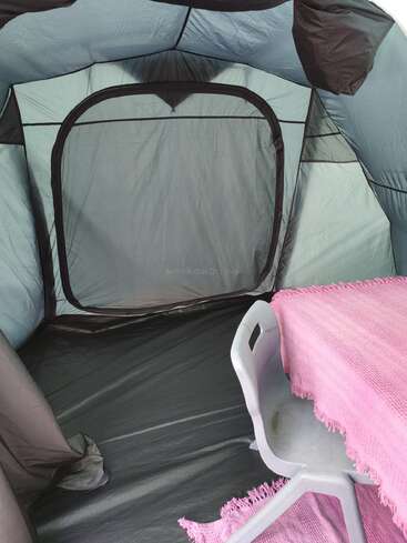 The image shows the inside of a tent with a mesh zippered door. There is a plastic chair and a table covered with a pink cloth.