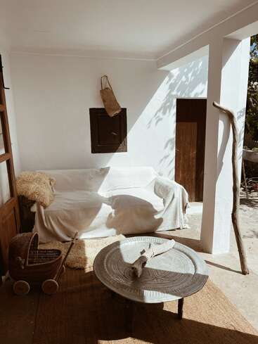 La imagen muestra una serena zona de estar al aire libre con un sofá blanco, una mesa de centro y una cesta de mimbre, rodeada por una puerta rústica de madera y un ambiente natural y terroso.