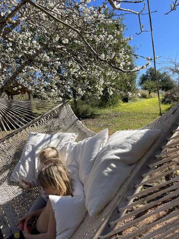 Una mujer de pelo rubio se sienta en una hamaca bajo un árbol en flor, rodeada de almohadas blancas y un sereno paisaje natural en un día soleado.