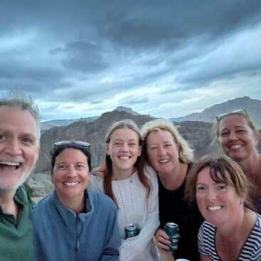 Six personnes souriantes prennent un selfie de groupe joyeux en plein air, avec un ciel nuageux et des montagnes pittoresques en arrière-plan. Ils semblent profiter d'un moment de détente ensemble.