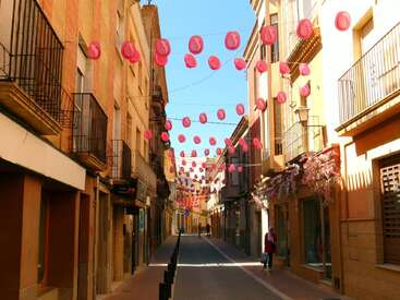A narrow street lined with colorful buildings has pink hats strung across overhead. Bright sunlight, blue sky, balconies, and a few pedestrians create a festive atmosphere.
