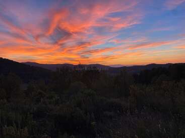 A stunning sunset paints the sky with vibrant orange, pink, and purple hues. Silhouetted hills and dark foliage create a peaceful and dramatic landscape scene.