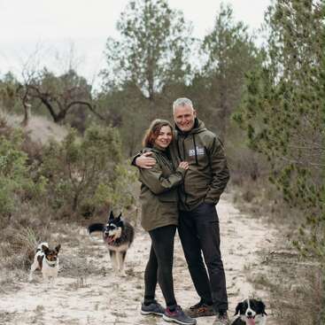 A couple stands together in a forest path, smiling and embracing, wearing matching jackets. Three dogs surround them, and green pine trees fill the background.