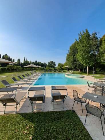 Une piscine d'un bleu limpide, entourée de chaises longues et de parasols, se trouve dans un jardin verdoyant, avec des arbres et un ciel lumineux qui créent une atmosphère paisible.