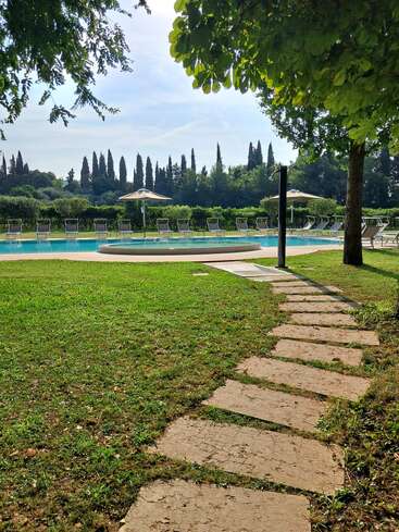 Un sentier en pierre traverse une pelouse et mène à une piscine extérieure tranquille, entourée de chaises longues, de parasols, d'une végétation luxuriante et de grands arbres en arrière-plan.