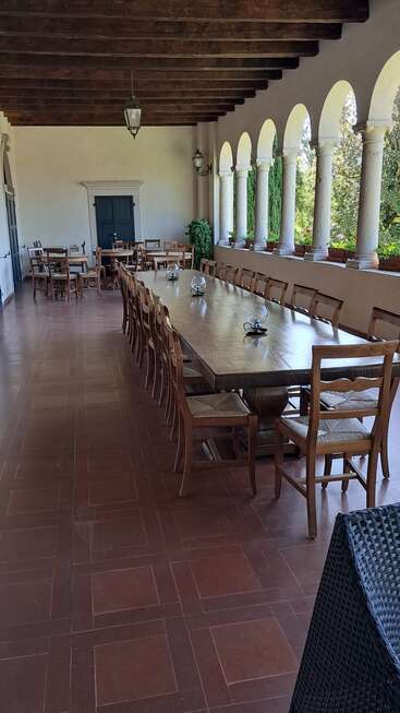Cette image représente une terrasse extérieure couverte avec une longue table à manger en bois, des chaises en bois, des colonnes arquées, un sol carrelé, des plantes en pot et un éclairage de type lanterne.